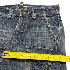 May include: Close-up of blue denim cargo pants with visible stitching and pockets. A yellow measuring tape shows a measurement of approximately 10 inches. The jeans have a button closure and metal rivets.