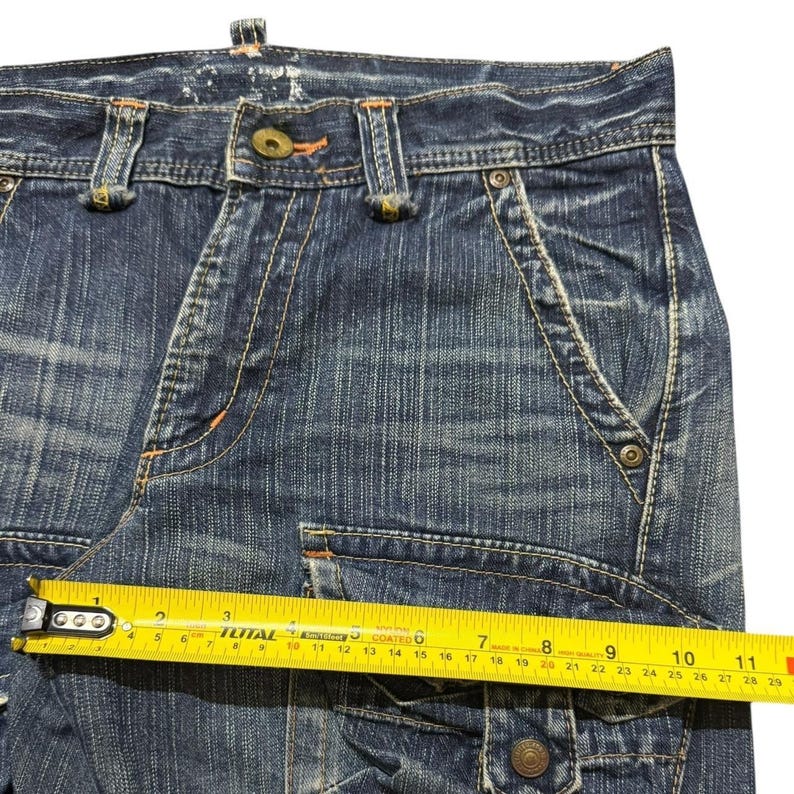 May include: Close-up of blue denim cargo pants with visible stitching and pockets. A yellow measuring tape shows a measurement of approximately 10 inches. The jeans have a button closure and metal rivets.