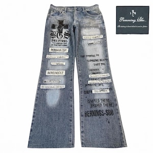 May include: Light blue denim jeans with a wide leg design. The jeans feature various printed text elements and patches. A black cross graphic is visible on the front. A small black label with text is also present.