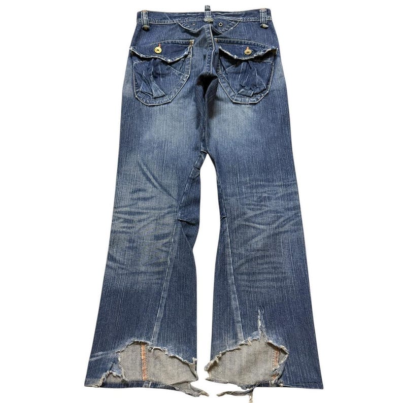 May include: Faded blue denim jeans with distressed details. The jeans feature unique pocket designs and flared legs with ripped hems. The back view showcases the intricate stitching and button details, offering a vintage aesthetic.