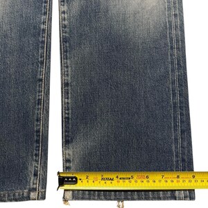 May include: A pair of blue denim jeans with a faded wash and straight leg cut. A yellow measuring tape is positioned across the bottom hem, showing a width of approximately 8.75 inches for the leg opening of the denim fabric.