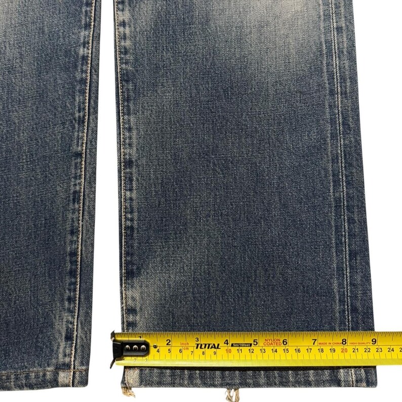 May include: A pair of blue denim jeans with a faded wash and straight leg cut. A yellow measuring tape is positioned across the bottom hem, showing a width of approximately 8.75 inches for the leg opening of the denim fabric.