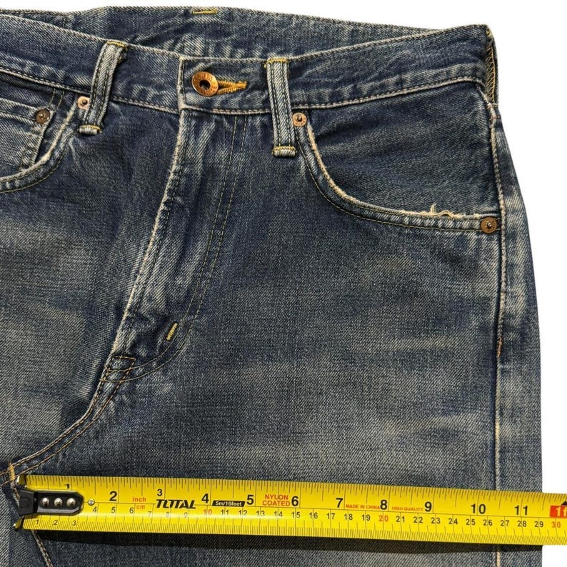 May include: Blue denim jeans featuring a medium wash, a button fly closure with a copper-colored button, and copper rivets at the pockets. A yellow measuring tape is positioned across the front, showing a width of approximately 11.5 inches.