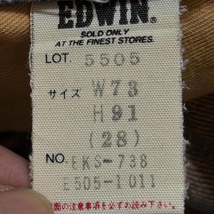 May include: A cream-colored fabric clothing tag with black text reading EDWIN, SOLD ONLY AT THE FINEST STORES, LOT 5505, SIZE W73 H91 (28), and NO. EKS-738 E505-1011. The tag is rectangular with frayed edges and is attached to a brown garment.