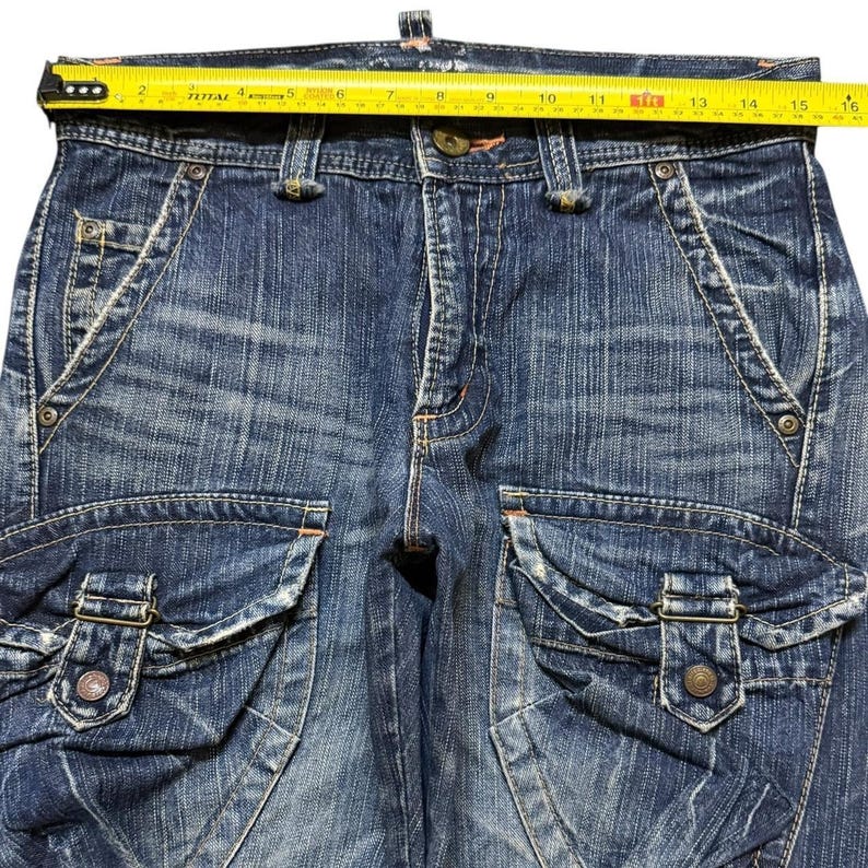 May include: Close-up of blue denim cargo pants with multiple pockets and metal button details. A yellow measuring tape is laid across the top of the pants, showing a measurement of approximately 16 inches.