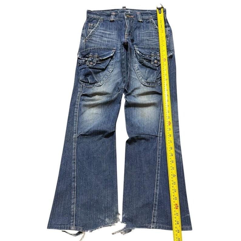 May include: Blue denim jeans with large cargo pockets on the thighs. The jeans have a wide leg and are measured with a yellow tape measure. The jeans are distressed at the bottom.