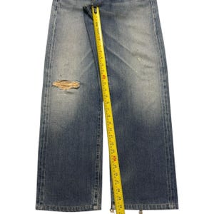 May include: Blue denim jeans featuring a distressed rip on the left leg. A yellow measuring tape is placed vertically along the center, showing a length measurement extending past 30 inches. The jeans have a faded wash and straight leg cut.
