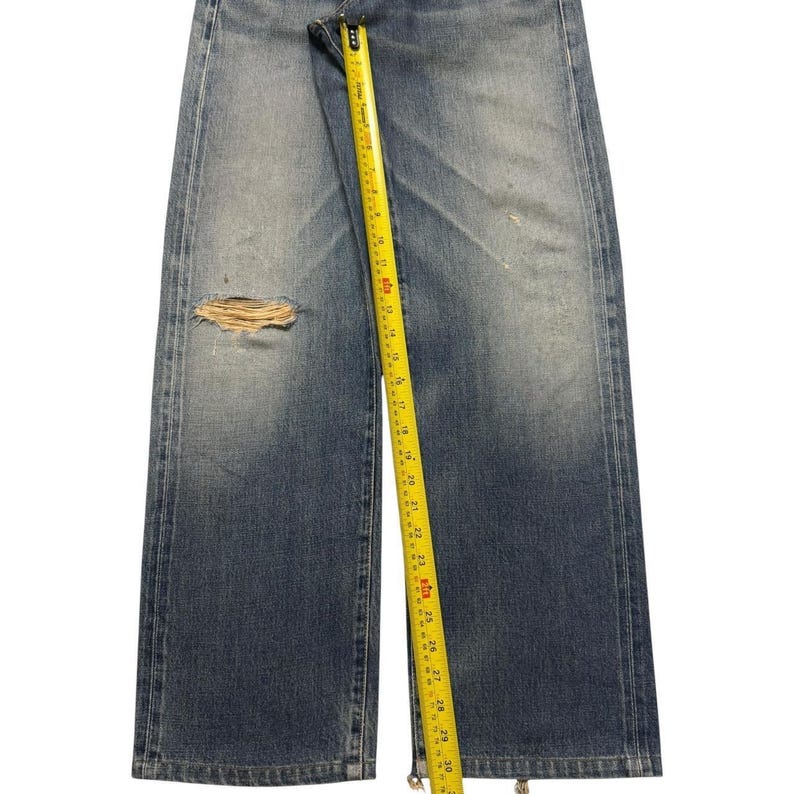 May include: Blue denim jeans featuring a distressed rip on the left leg. A yellow measuring tape is placed vertically along the center, showing a length measurement extending past 30 inches. The jeans have a faded wash and straight leg cut.
