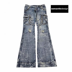 Puede incluir: Pantalones cargo de mezclilla azul con detalles desgastados, con múltiples bolsillos y cremalleras. Los pantalones tienen un diseño de pierna acampanada y el nombre de la marca "semanticdesign" es visible en un rectángulo negro.