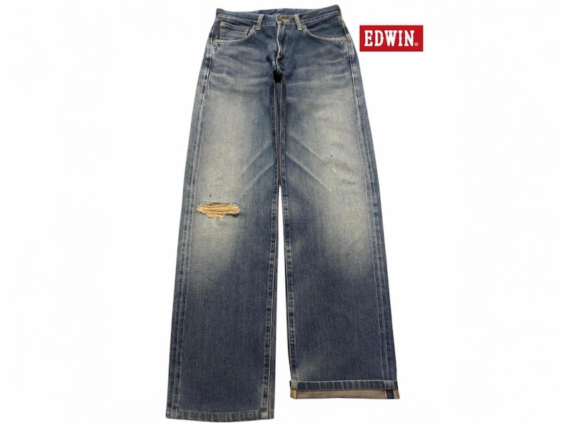 May include: Blue denim jeans by EDWIN featuring a faded wash, a straight-leg silhouette, and a distressed rip on the right thigh. The jeans include a button fly, belt loops, and classic five-pocket styling with visible whiskering and fading.
