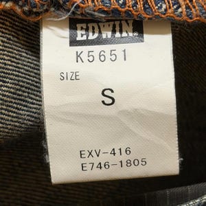 May include: Close-up of a white tag with the brand name "EDWIN" and the code "K5651" printed in black. The tag also displays "SIZE S" and the codes "EXV-416" and "E746-1805". The tag is attached to denim fabric.
