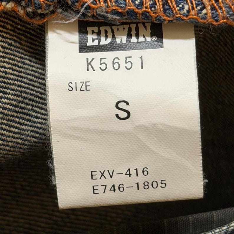 May include: Close-up of a white tag with the brand name "EDWIN" and the code "K5651" printed in black. The tag also displays "SIZE S" and the codes "EXV-416" and "E746-1805". The tag is attached to denim fabric.