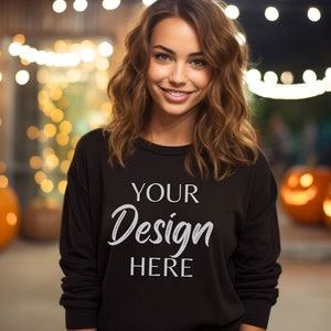 Könnte beinhalten: Ein schwarzes Sweatshirt mit dem Text "YOUR Design HERE" in weißer Schrift. Das Bild ist im Freien bei Nacht aufgenommen, mit Lichterketten und geschnitzten Kürbissen im Hintergrund, was auf ein Halloween-Thema hindeutet.