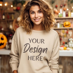 Könnte beinhalten: Beiges Sweatshirt mit dem Text "YOUR Design HERE" in Dunkelgrau. Die Person lächelt, mit einem verschwommenen Hintergrund mit Halloween-Dekorationen und warmer Beleuchtung.