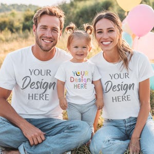 Può includere: Una famiglia di tre persone indossa magliette bianche abbinate con la scritta "YOUR Design HERE". La maglietta del bambino ha un design colorato e la scritta "YOUR Design HERE". Sono seduti sull'erba con palloncini sullo sfondo.