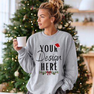 Pode incluir: Um moletom cinza claro com o texto "YOUR Design HERE" em escrita preta, um gorro de Papai Noel vermelho e um desenho de azevinho e laço. A pessoa está segurando uma caneca branca em frente a uma árvore de Natal.