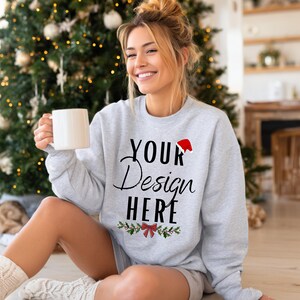 Pode incluir: Um moletom cinza claro com o texto "YOUR Design HERE" em escrita preta e um gorro de Papai Noel vermelho. O moletom tem um tema de Natal com um design de azevinho e laço. Uma caneca branca é segurada na mão da pessoa.