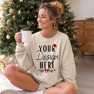 Könnte beinhalten: Ein cremefarbenes Sweatshirt mit dem Text "YOUR Design HERE" in schwarzer Schrift, einer roten Weihnachtsmütze und festlichem Grün. Die Person hält einen weißen Becher, mit einem Weihnachtsbaum im Hintergrund.