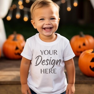 Könnte beinhalten: Ein kleines Kind trägt ein weißes T-Shirt mit dem Aufdruck "YOUR Design HERE". Das Kind lächelt und steht vor einem Hintergrund aus Kürbissen.