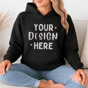 K&ouml;nnte beinhalten: Schwarzer Kapuzenpullover mit dem wei&szlig;en Text "YOUR DESIGN HERE". Der Pullover hat eine Vordertasche und eine Kapuze mit Kordelzug. Das Model tr&auml;gt eine blaue Jeans.