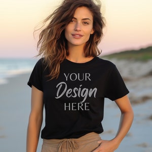 Puede incluir: Camiseta negra de cuello redondo con el texto "YOUR Design HERE" en fuente cursiva. La modelo lleva pantalones color caqui y está de pie en una playa. El fondo muestra el mar y una puesta de sol.