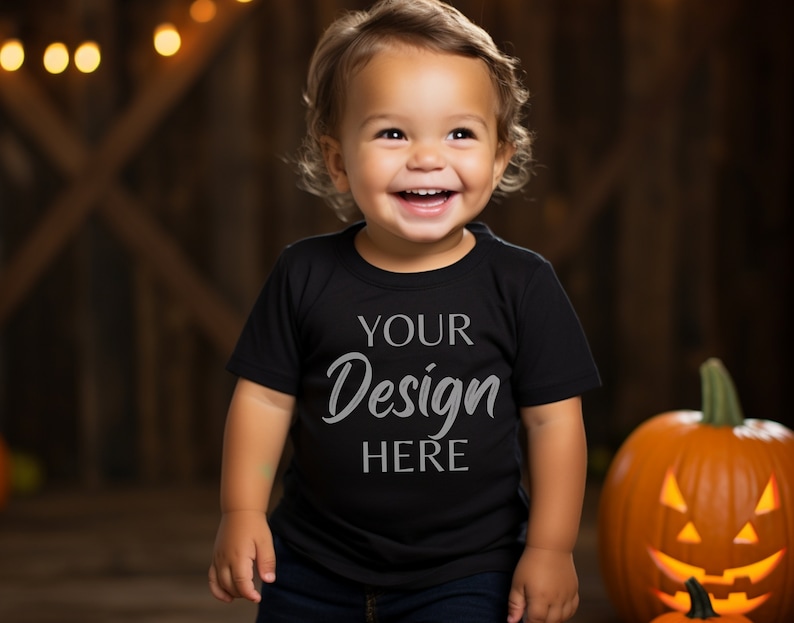 K&ouml;nnte beinhalten: Ein l&auml;chelndes Kleinkind tr&auml;gt ein schwarzes T-Shirt mit dem Text "YOUR Design HERE". Der Hintergrund umfasst einen geschnitzten K&uuml;rbis und eine rustikale Holzwand. Warme Lichter sind im Hintergrund aufgereiht.