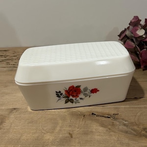 Peut inclure: Un porte-savon rectangulaire blanc avec un couvercle à carreaux et un motif floral de roses. Le plat est fait d'un matériau brillant et repose sur une surface en bois. Un bouquet de fleurs bordeaux séchées est en arrière-plan.