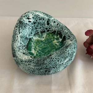 May include: A unique, handcrafted ceramic bowl with a textured, porous exterior in shades of teal, black, and white. The interior features a vibrant green, glossy finish. The bowl has an organic, asymmetrical shape, perfect for decorative use.