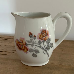 Peut inclure: Crémier en céramique blanche avec un motif floral de roses aux tons jaune, orange et rose. Le crémier a une poignée incurvée et un bord doré. Le motif floral est sur le devant.