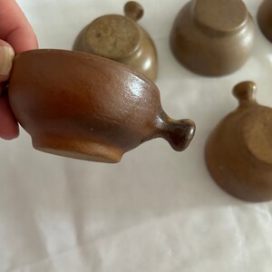 Puede incluir: Colección de pequeños cuencos de cerámica marrón con asa. Los cuencos tienen forma redondeada y un acabado liso y vidriado. El asa es un pequeño pomo redondeado. Los cuencos están dispuestos sobre una superficie blanca.