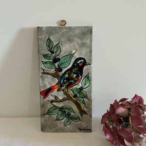 Può includere: Piastrella in ceramica rettangolare raffigurante un uccello colorato posato su un ramo con foglie verdi e bacche blu. L'uccello ha piume rosse, arancioni, nere e bianche. La piastrella ha uno sfondo grigio e un anello dorato per appenderla.