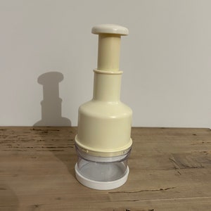 Puede incluir: Un prensa ajos de plástico blanco con un recipiente de plástico transparente. El prensa ajos tiene un émbolo que presiona hacia abajo sobre el ajo para machacarlo.