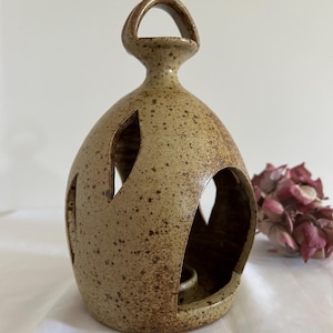 May include: A speckled ceramic candle holder with a handle and cut-out designs. The beige-coloured holder has brown speckles and a small opening to hold a candle. The design features a rounded shape with a handle at the top.