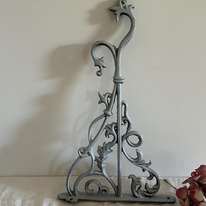 Peut inclure: Une équerre murale en fonte gris clair présentant un design orné et volute avec des motifs en forme de feuilles. Le support possède une barre verticale, un crochet supérieur incurvé et une base plate avec des trous de vis visibles.