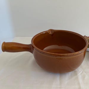 Puede incluir: Un cuenco de sopa de cerámica marrón con asa. El cuenco tiene forma redondeada y acabado brillante. El asa está fijada al lateral del cuenco. Hay flores secas junto al cuenco.