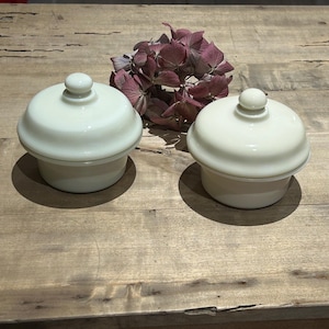 May include: Two matching off-white custard cups with rounded lids and small spherical knobs. Each cup features a smooth, opaque finish and a wide, slightly flared rim design. The containers are circular with a clean, simple silhouette.