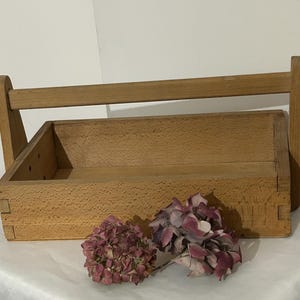 Könnte beinhalten: Eine hellbraune Werkzeugkiste aus Holz mit einem Griff, mit einer rechteckigen Kastenform. Die Kiste ist mit sichtbaren Verbindungen konstruiert und wird von getrockneten rosa Blumen begleitet. Die Werkzeugkiste ist für den Transport von Werkzeugen und Zubehör konzipiert.