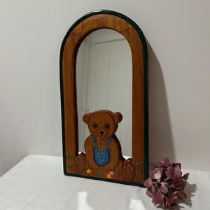 Puede incluir: Espejo enmarcado en madera con un diseño de oso de peluche marrón. El oso lleva un peto azul y está sentado en un campo de flores. El marco tiene forma de arco en la parte superior y tiene un borde verde.