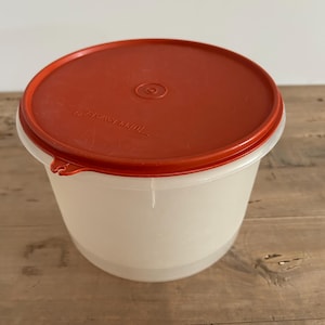 Könnte beinhalten: Ein durchsichtiger Plastikbehälter mit einem leuchtend orangenen Deckel. Der Deckel hat einen kleinen Kreis in der Mitte und der Text "Tupperware" ist auf dem Deckel aufgedruckt.