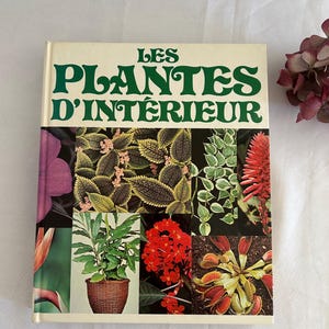 Puede incluir: Un libro vintage titulado "LES PLANTES D'INTÉRIEUR" con una cubierta color crema y letras verdes. La portada presenta un collage de coloridas plantas de interior. Un arreglo floral burdeos seco está en el fondo.