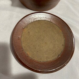 K&ouml;nnte beinhalten: Ein brauner Keramikdeckel f&uuml;r einen Tontopf. Der Deckel hat eine braune Glasur und einen erh&ouml;hten Rand. Die Unterseite des Deckels hat einen kreisf&ouml;rmigen, unglasierten Bereich mit der Zahl "2" darauf.