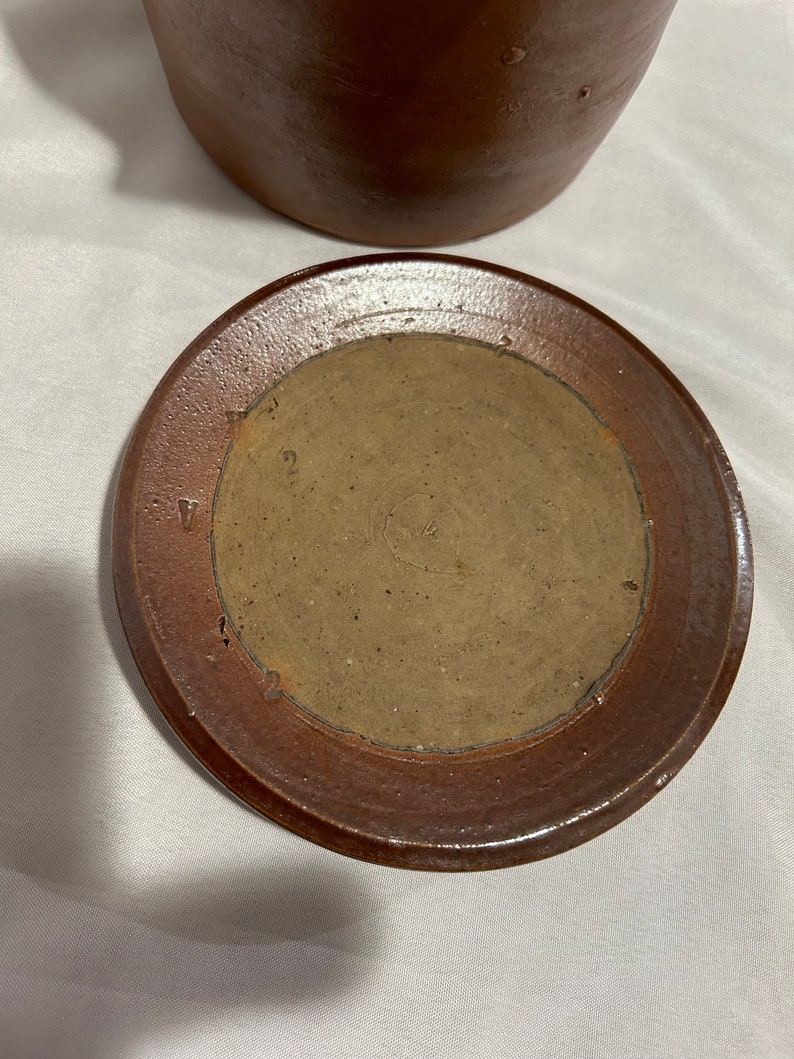 K&ouml;nnte beinhalten: Ein brauner Keramikdeckel f&uuml;r einen Tontopf. Der Deckel hat eine braune Glasur und einen erh&ouml;hten Rand. Die Unterseite des Deckels hat einen kreisf&ouml;rmigen, unglasierten Bereich mit der Zahl "2" darauf.