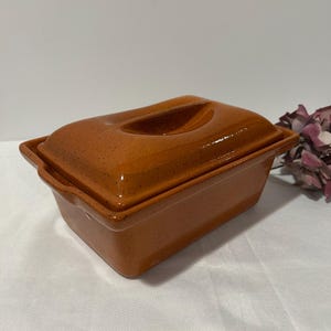 May include: An orange ceramic loaf pan with a matching lid. The pan has a rectangular shape with rounded edges and a speckled finish. The lid features a curved handle. The pan is on a white surface.