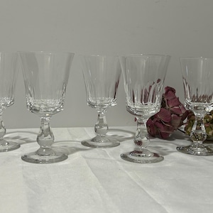 May include: Five clear crystal wine glasses with a decorative stem and a textured bowl. The glasses are arranged on a white surface, with dried flowers in the background. The glasses are ideal for serving wine.
