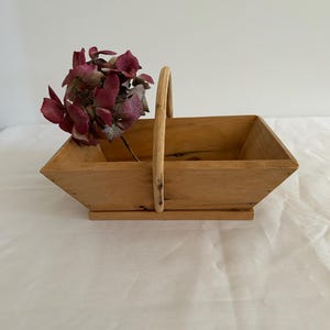 Könnte beinhalten: Ein Holzkorb mit Henkel, aus Naturholz mit hellbrauner Oberfläche. Der Korb ist leer, aber im Hintergrund befindet sich ein Strauß getrockneter lila Blumen.