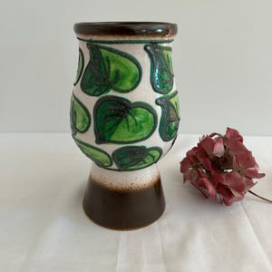 Puede incluir: Un jarrón de cerámica con base blanca y borde y base marrón. El jarrón está decorado con motivos de hojas verdes. Una flor seca con pétalos rojos está al lado del jarrón.