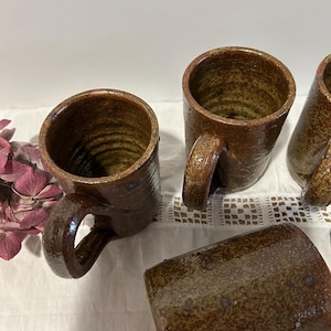 May include: A set of brown ceramic mugs with handles, showcasing a rustic, textured finish. The mugs display colour variations and a glazed appearance. The interior of the mugs has a spiral pattern. The mugs are placed on a white surface.