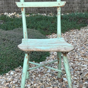 Peut inclure: Une chaise en bois vert clair vieilli au design simple. La chaise a un dossier et un siège à lattes, et les pieds sont légèrement évasés. La chaise est posée sur un lit de petites pierres.