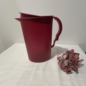 以下が含まれることがあります： ブルゴーニュ色のプラスチック製ピッチャー。蓋とハンドルが付いています。ピッチャーは空で、白い表面に置かれ、乾燥した花の小さな束の隣にあります。ピッチャーは飲み物を提供するように設計されています。