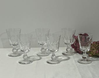 May include: Set of six clear crystal wine glasses. Each glass features a decorative, textured pattern on the bowl and a round base. The glasses are arranged on a white surface, with a few pink flowers in the background.